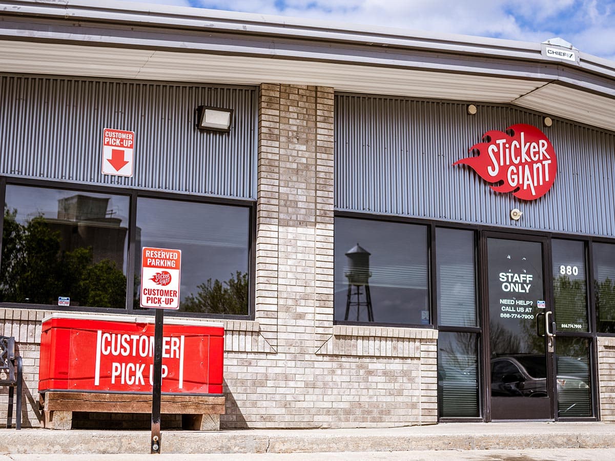 The exterior of a StickerGiant building shows a red StickerGiant sign above the entrance and a red customer pickup box near the front windows.