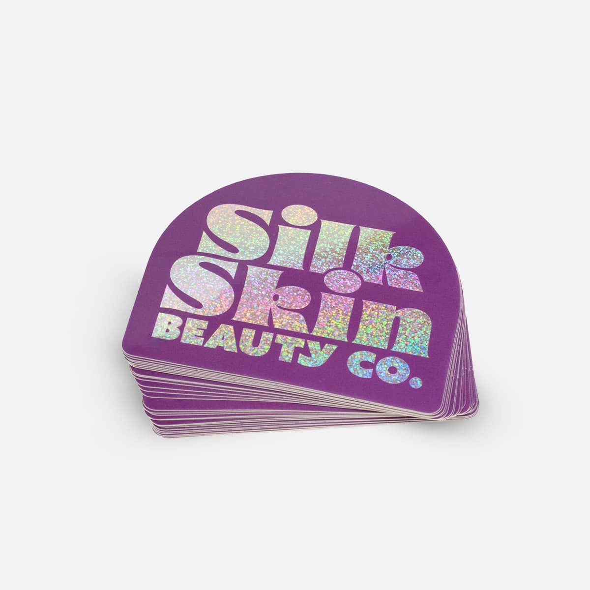 Stack of custom glitter stickers with holographic lettering for Silk Skin Beauty Co. on a purple background.