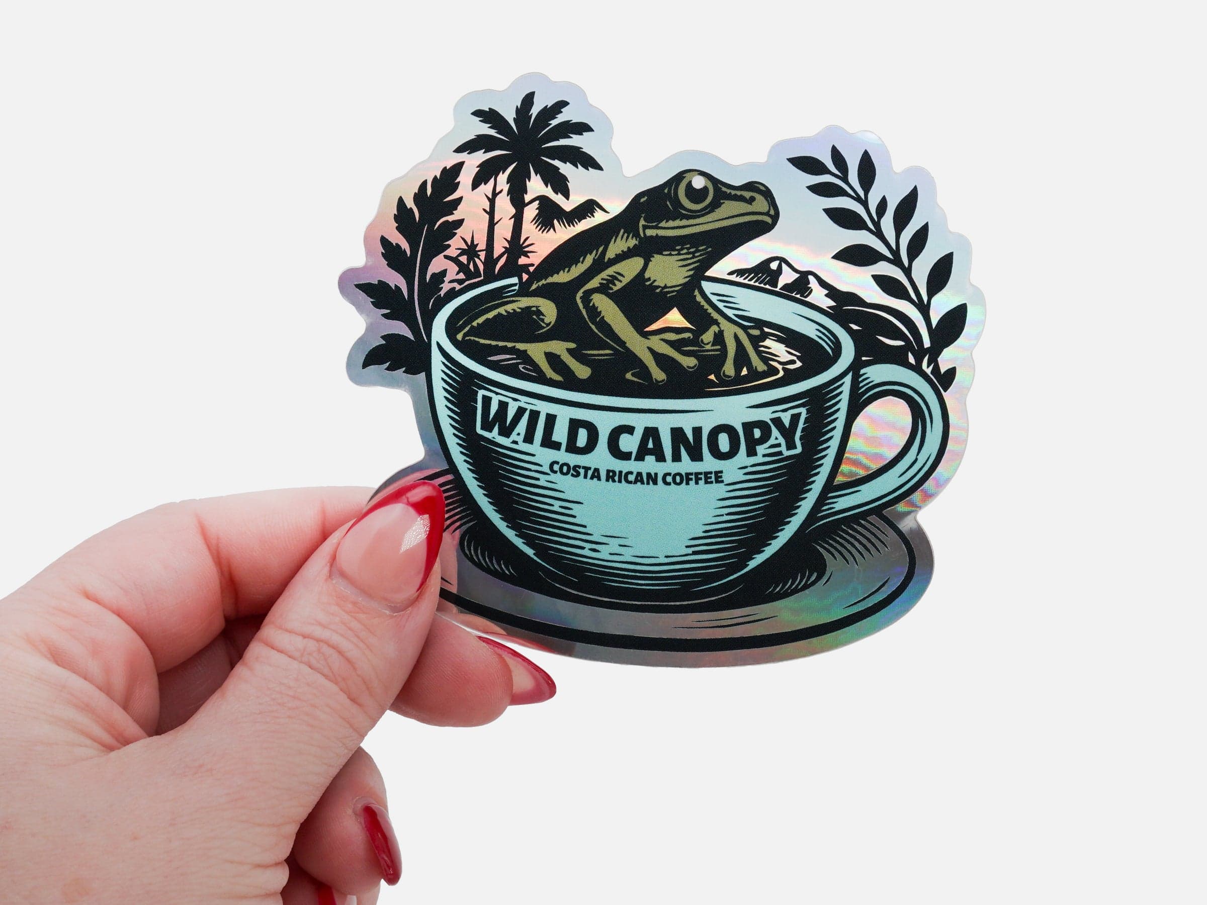 Holographic die cut coffee sticker featuring a frog sitting in a coffee cup with tropical foliage, held in a person's hand.