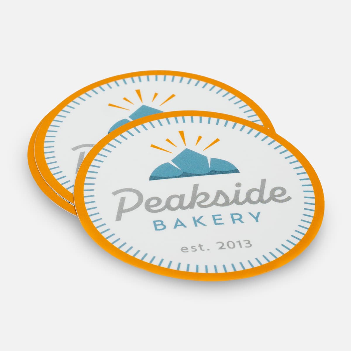 Stack of oval Peakside Bakery stickers with an orange border, blue mountain icon, and “est. 2013” text.