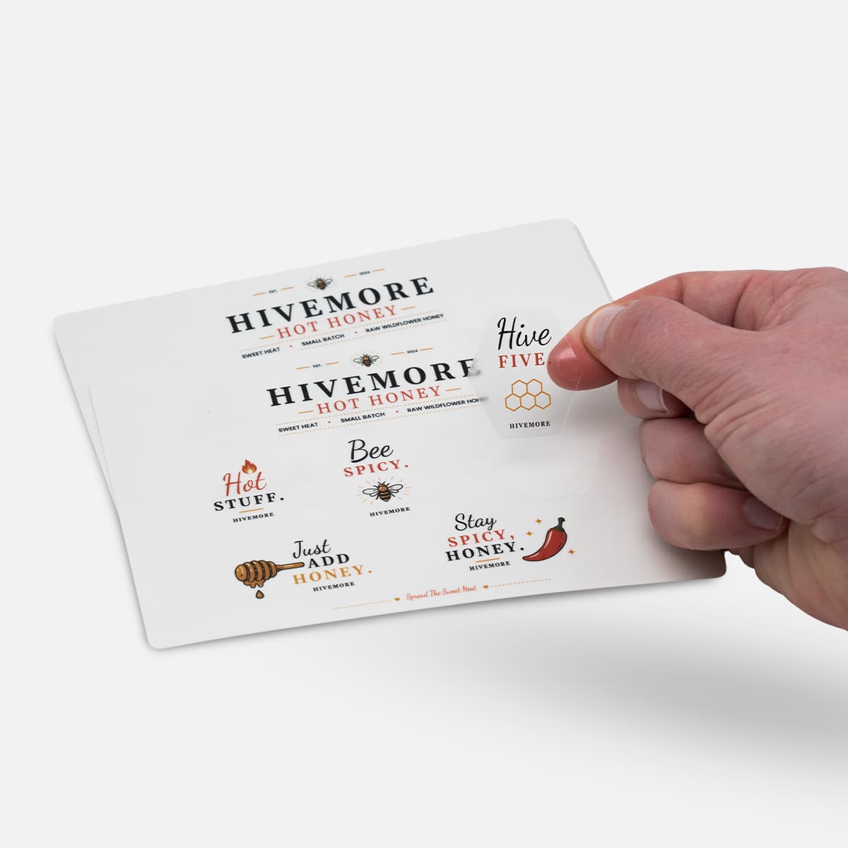 Custom Hivemore Hot Honey sticker sheet with multiple spicy honey designs, including a clear Hive Five sticker being peeled off by hand.