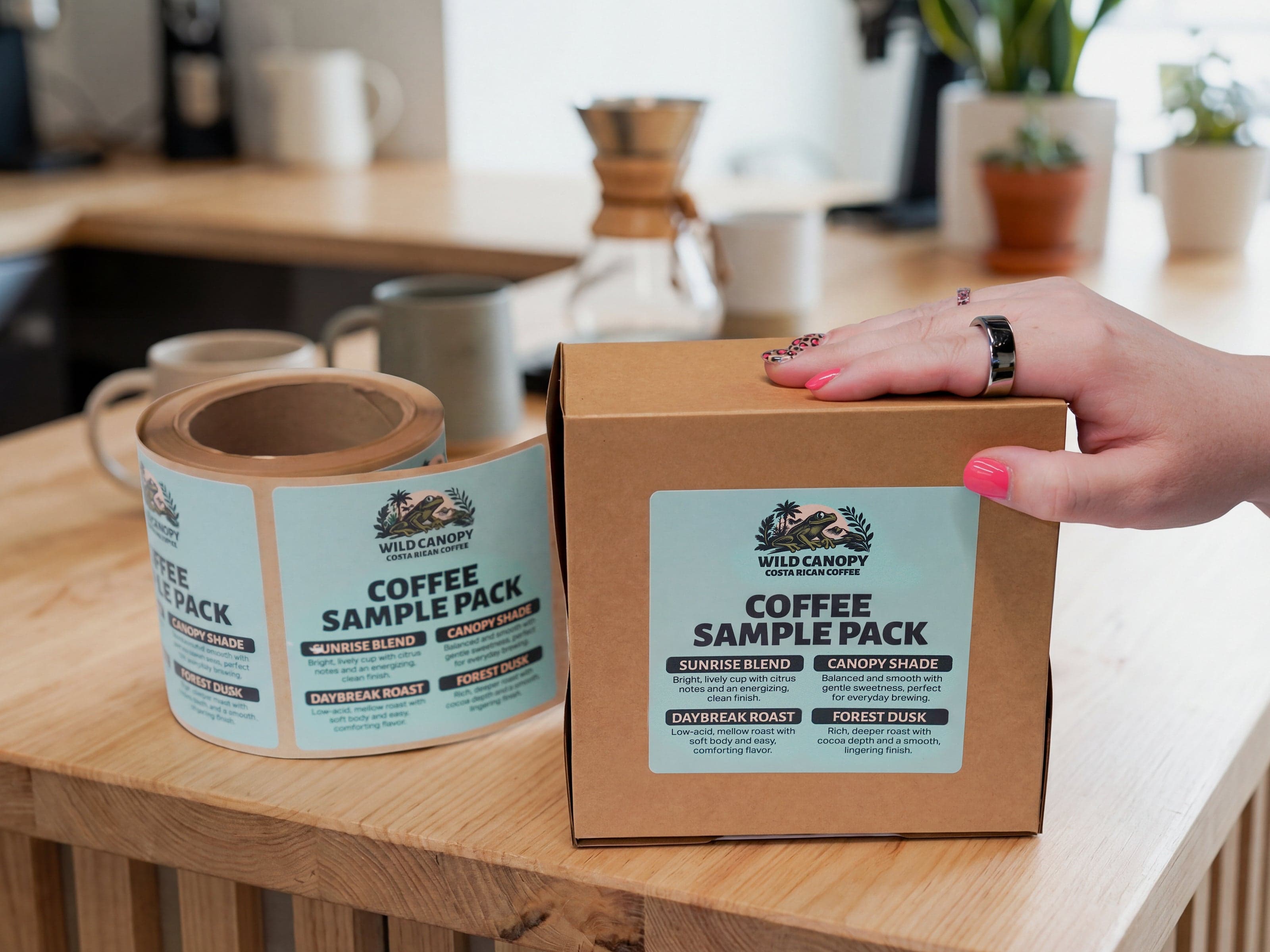 Roll of custom coffee product labels next to a labeled coffee sample pack box on a tabletop in a coffee shop setting.