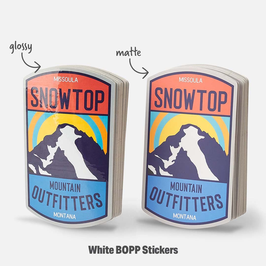 Stacks of custom white BOPP stickers featuring a mountain outfitter design, shown side by side to compare glossy and matte laminate finishes.