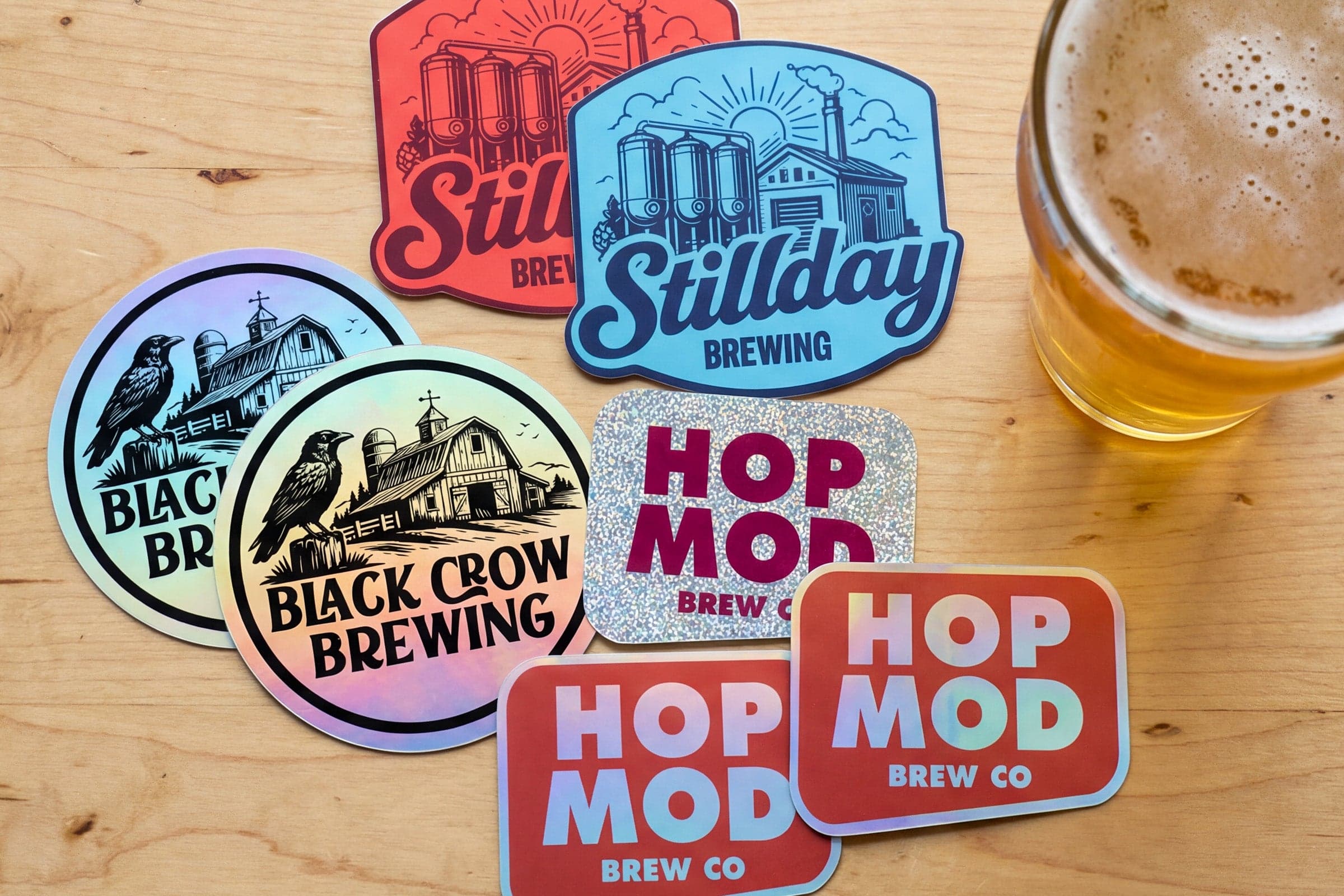 Custom brewery stickers in holographic, glitter, and matte finishes displayed on a tabletop next to a glass of beer, showcasing branded craft beer promotional stickers.