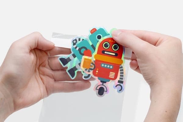 Hands placing colorful robot stickers into a clear plastic sticker pack bag.