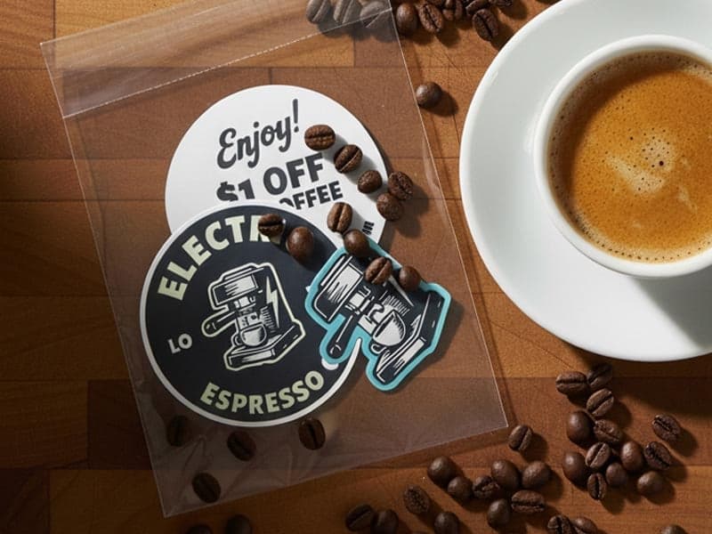Coffee shop sticker pack with branded coffee stickers and coupon card inside a clear resealable bag next to a cup of espresso.